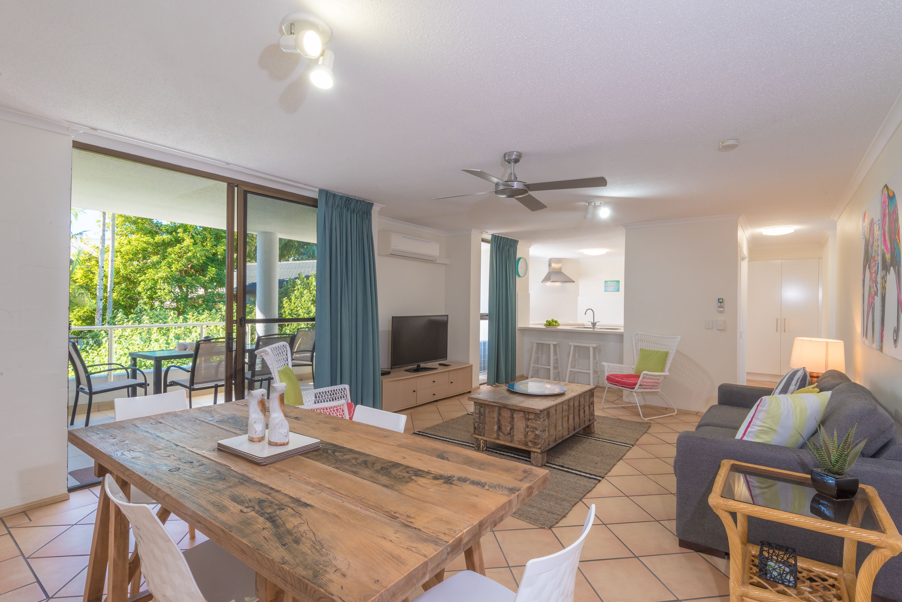 Un salon moderne avec une table à manger en bois, un canapé et un téléviseur. On aperçoit un balcon avec des chaises et une table, ainsi qu’un coin cuisine au fond.