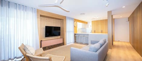 Un salon moderne avec un canapé, un fauteuil, un téléviseur à écran plat et un coin cuisine à l’arrière-plan.