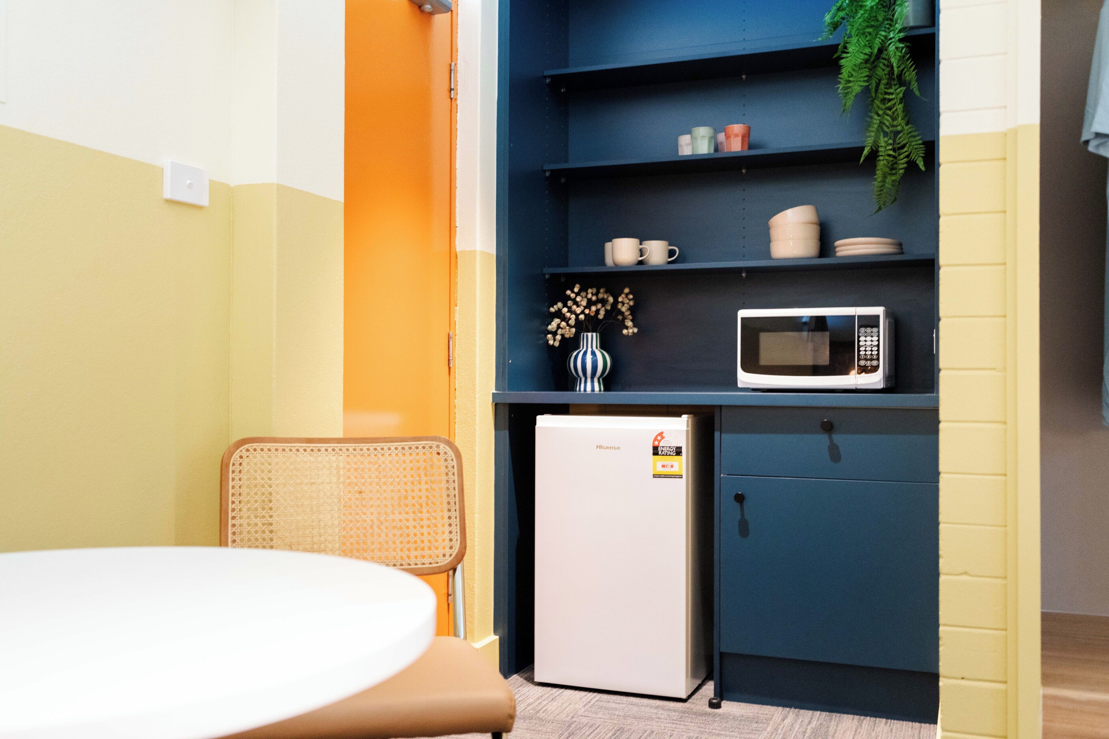 Una kitchenette compacta con microondas, un refrigerador pequeño y una alacena con tazas y un florero.