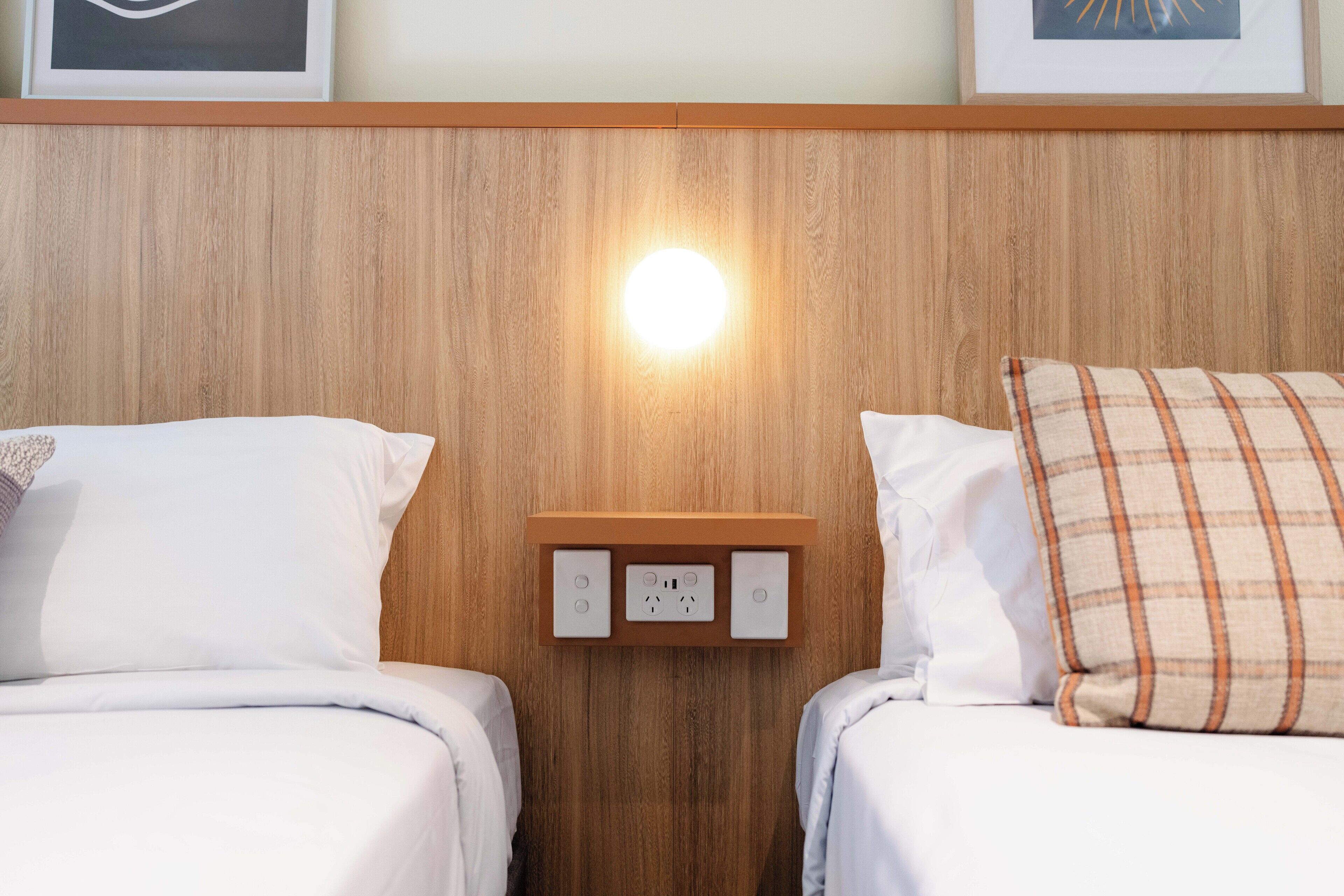 Hotellihuone, jossa on kaksi sänkyä, puinen pääty ja seinään kiinnitetty valaisin sekä pistorasia.