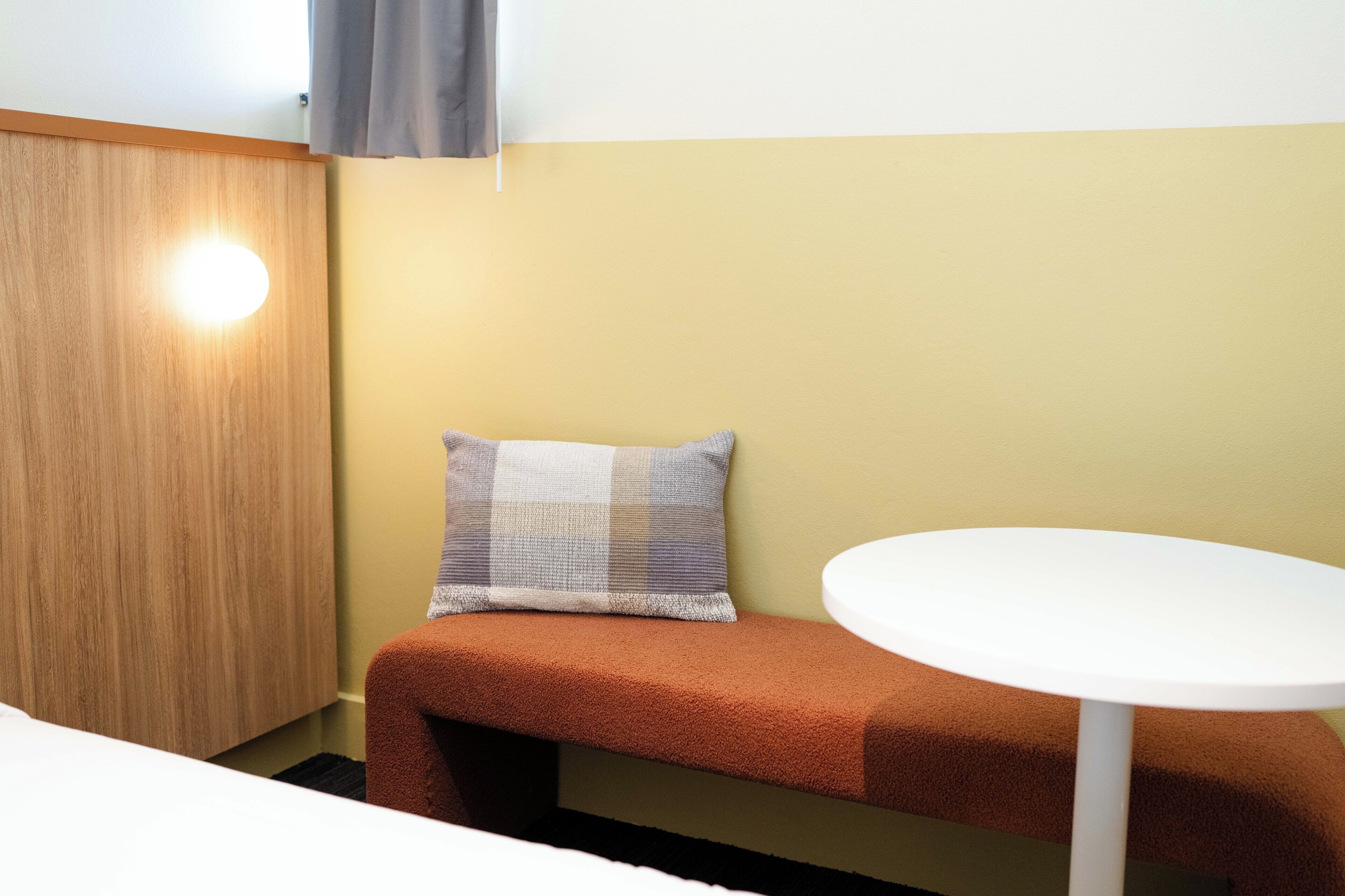 A small room with a wooden wardrobe, a bench with a cushion, and a round white table.