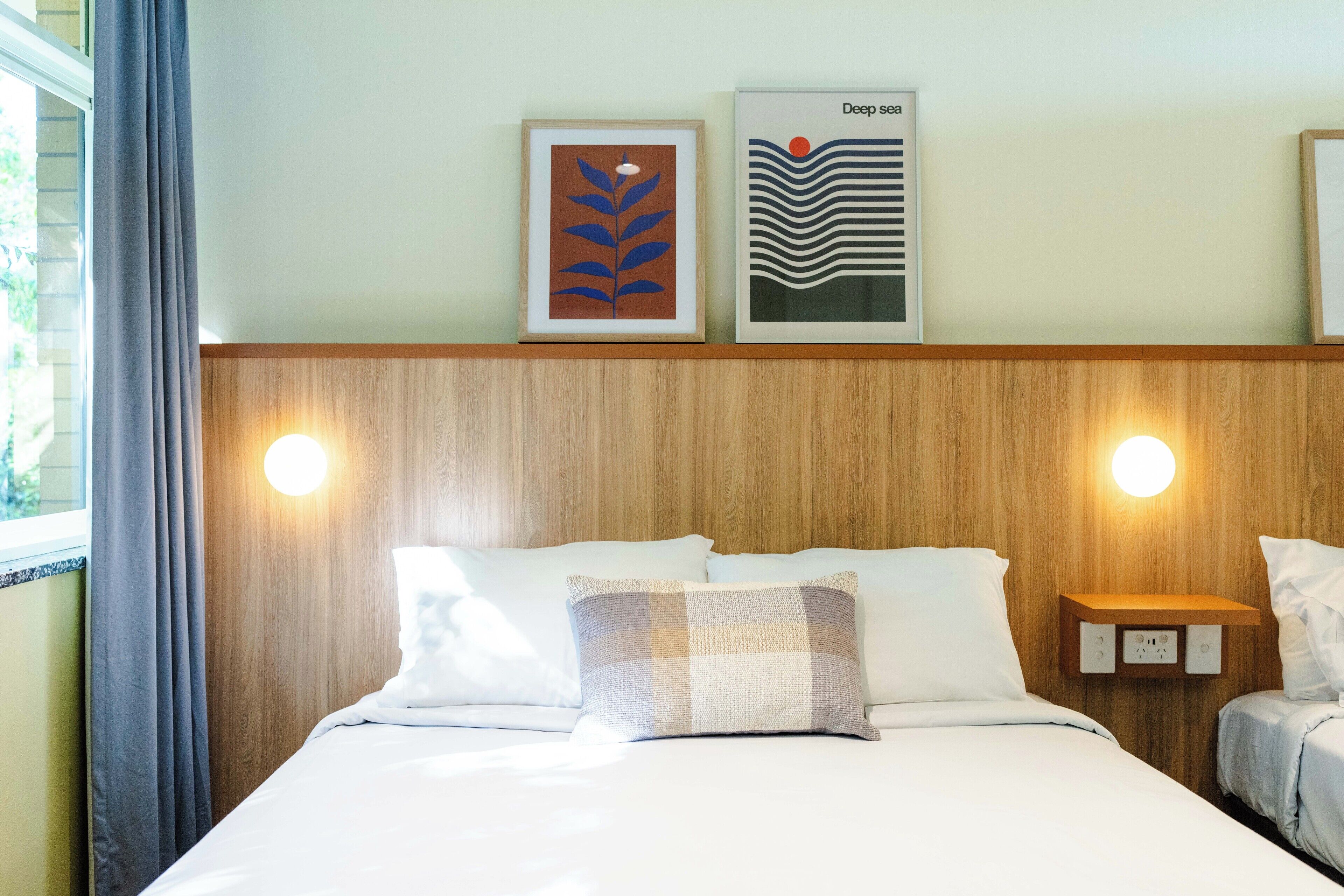 Een hotelkamer met een houten hoofdeinde, twee bedden, een nachtkastje met een lamp en twee ingelijste kunstwerken aan de muur.
