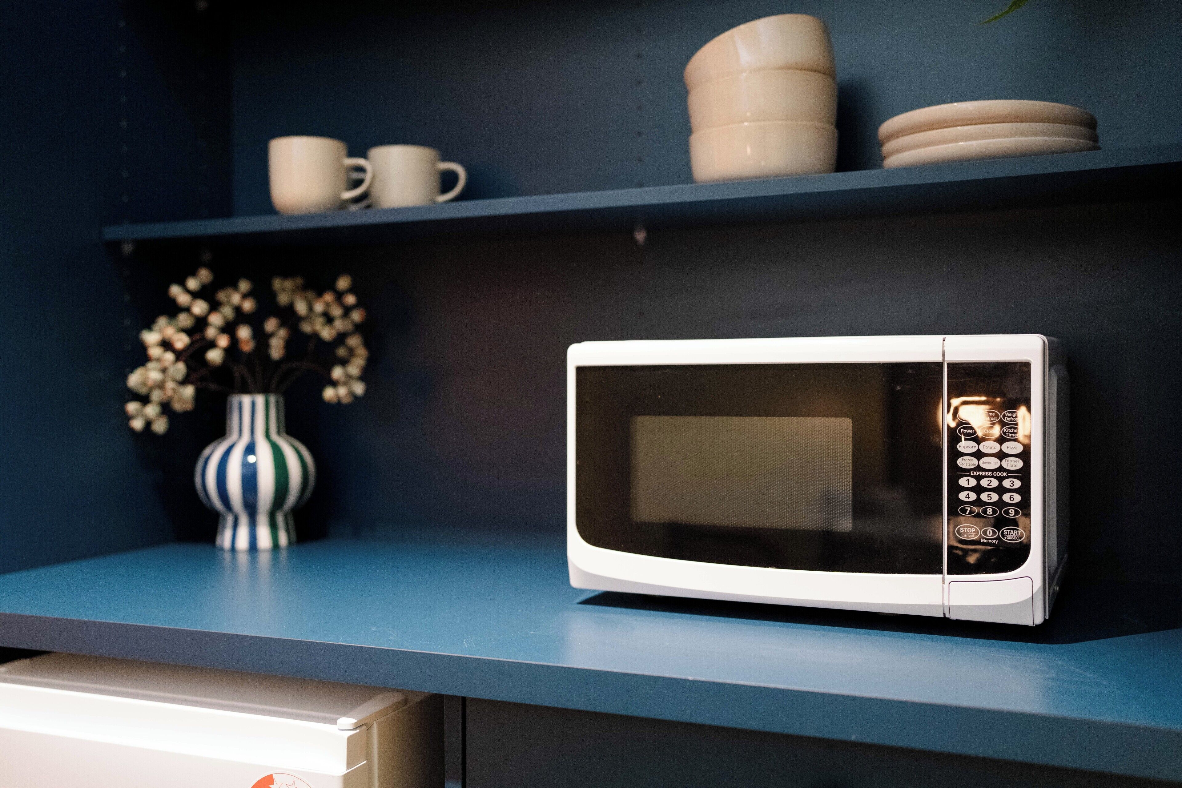 A microwave on a blue countertop with a vase of flowers and stacked plates on a shelf above.
