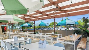 Café al aire libre con mesas, sillas, sombrillas y vistas a coches estacionados y follaje.