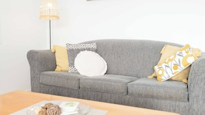 Un sofá gris con cojines estampados, una bandeja de cristal transparente con objetos decorativos y una lámpara de pie tejida.