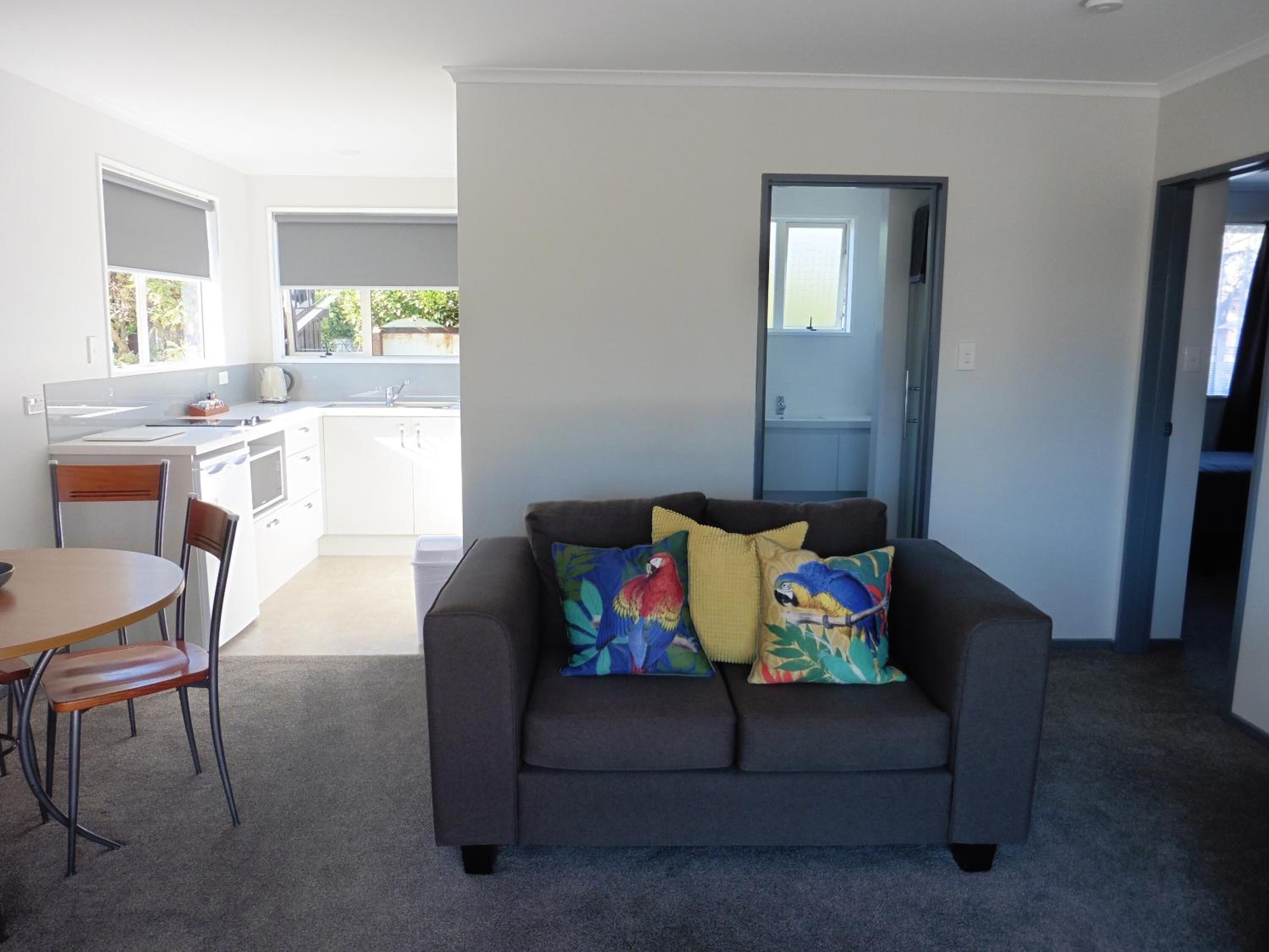 A modern living room with a grey sofa, a wooden dining table, and chairs. There is a kitchen area with white cabinets and a window.
