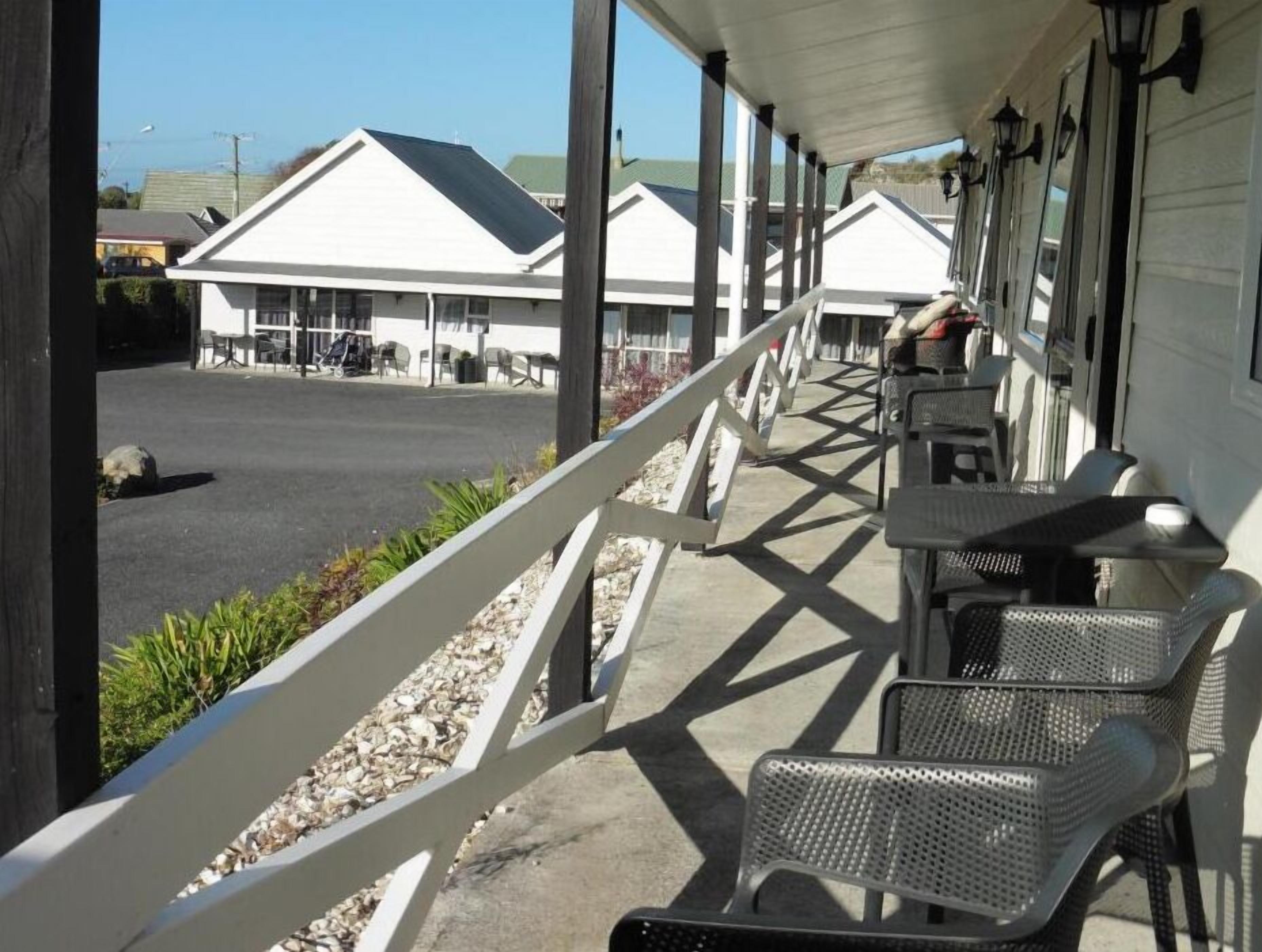 A covered outdoor seating area with metal chairs and tables, a white railing, and a view of a building with a white facade and a green roof.