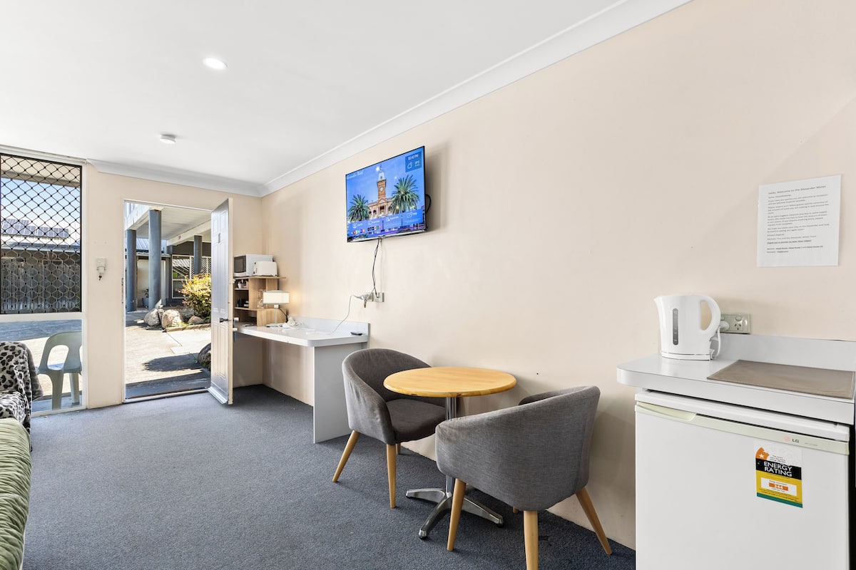 A modern hotel room with a flat-screen TV mounted on the wall, a small round table with two chairs, a kitchenette area, and a view through a glass door to an outdoor area.