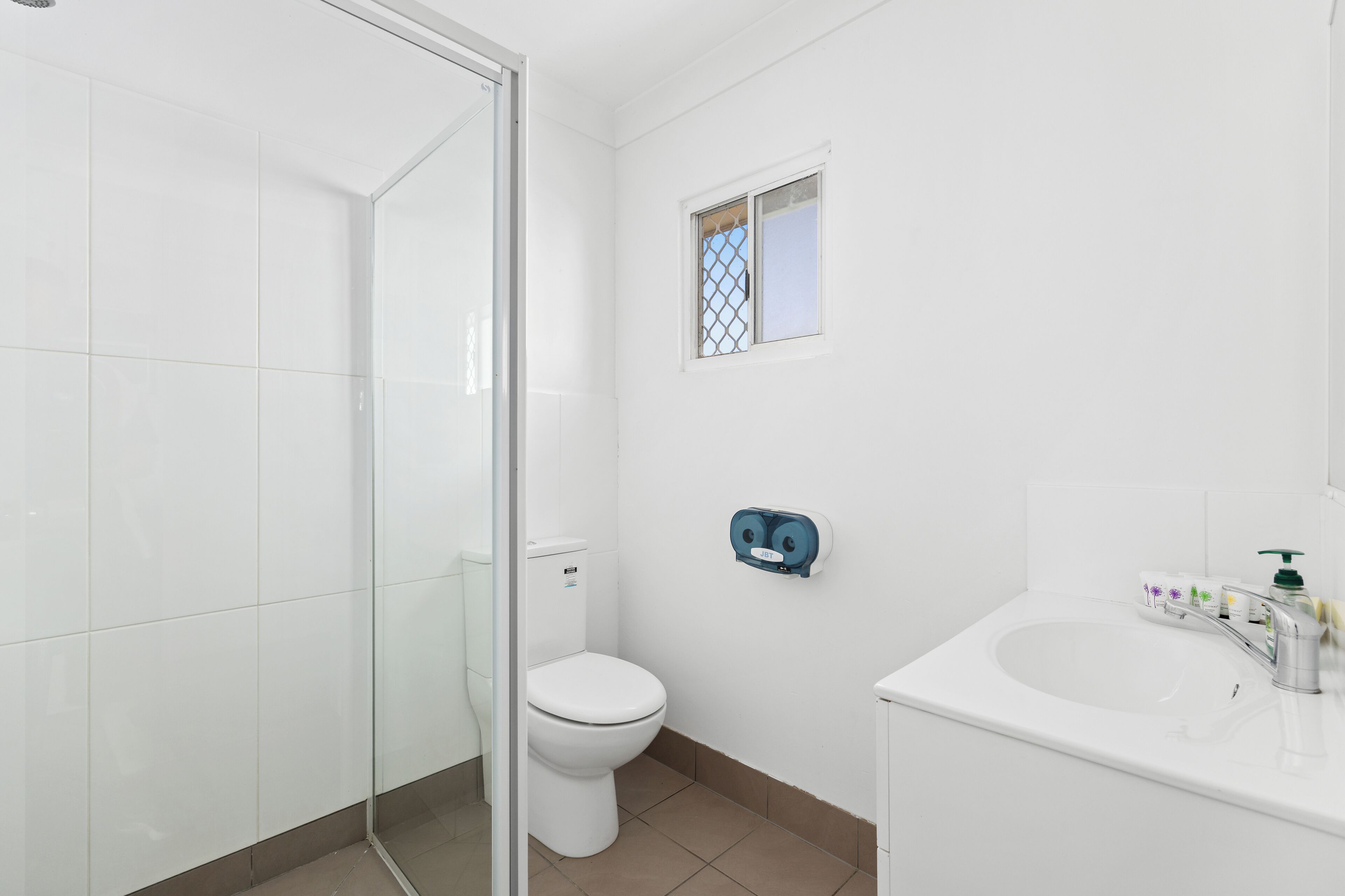 Ein modernes Badezimmer mit Duschkabine aus Glas, Toilette, kleinem Fenster und Waschbecken mit Pflegeprodukten.