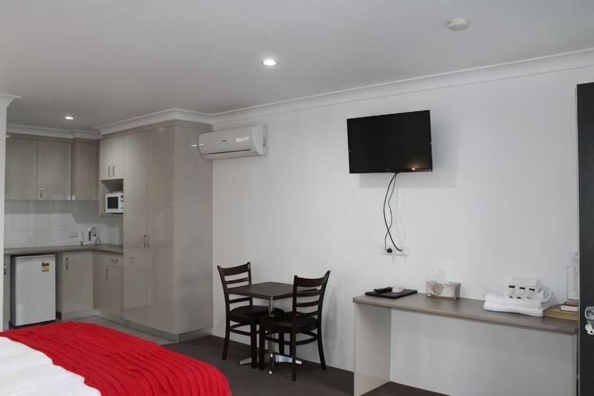 A compact hotel room with a kitchenette, a bed with a red blanket, a TV mounted on the wall, and a small dining table.