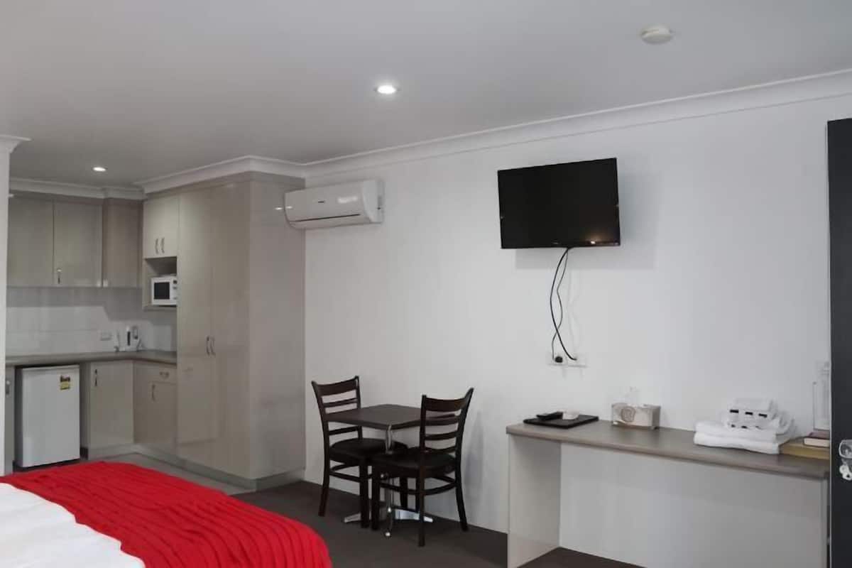Habitación de hotel compacta con kitchenette, una cama con una manta roja, un televisor montado en la pared y una pequeña mesa de comedor.