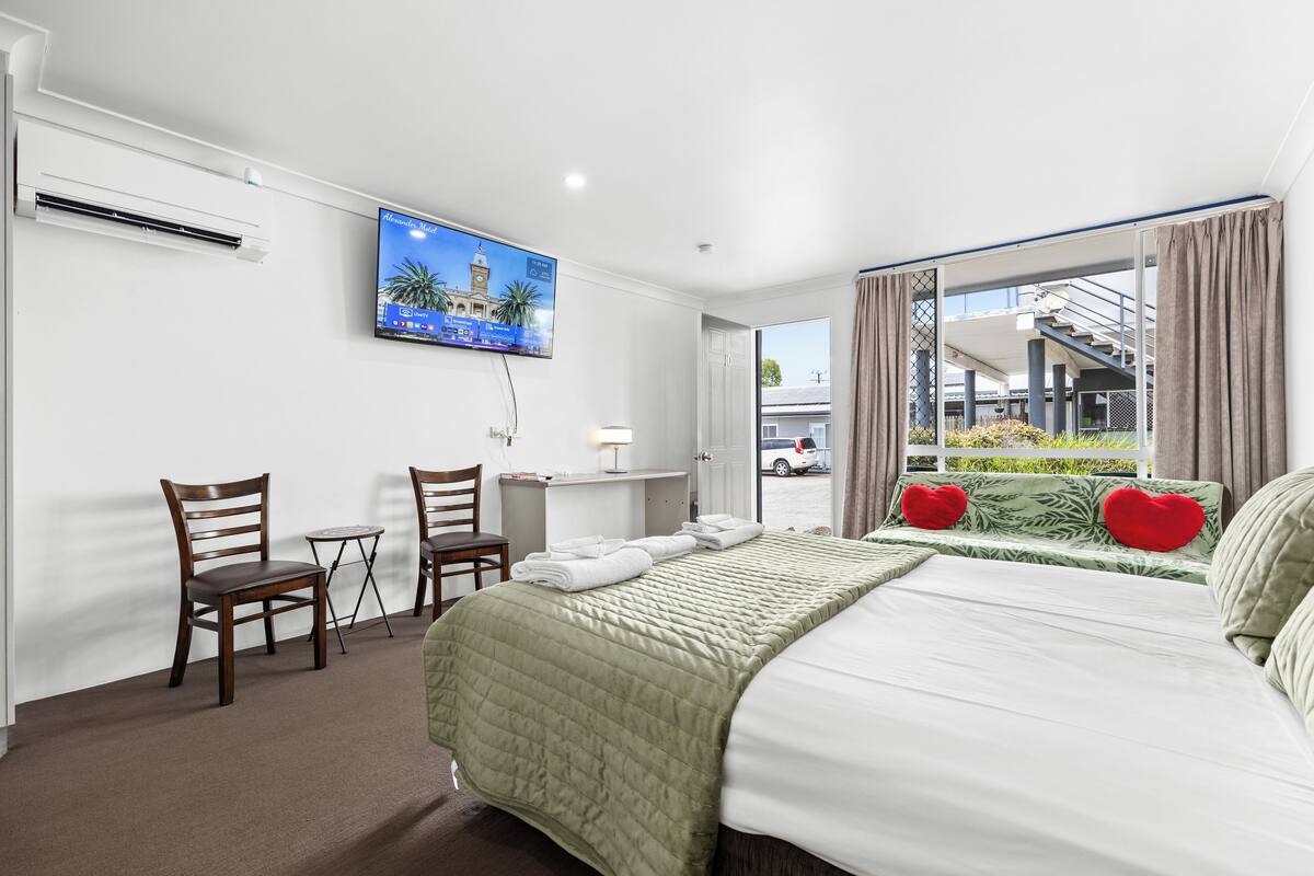 A hotel room with a bed, two chairs, a small table, a TV on the wall, and a window with curtains.