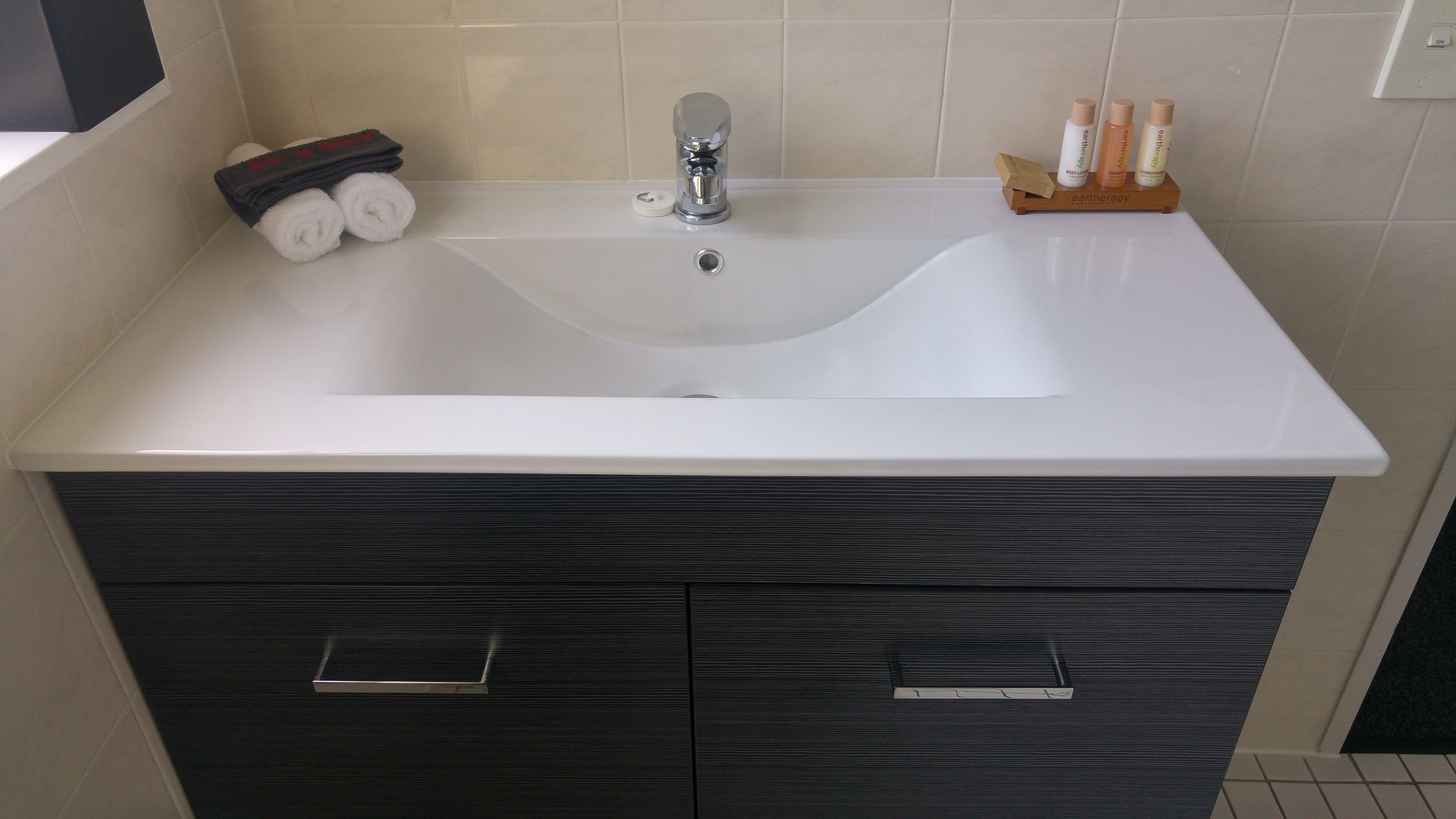 Un baño con lavamanos blanco, muebles de madera oscura y un pequeño estante con artículos de aseo.