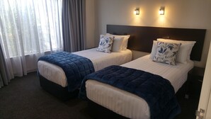 A hotel room with two beds, each with a dark blue quilted bedspread and white linens. There are two wall-mounted light fixtures above the beds, and a window with sheer curtains to the left. - White Heron Motor Lodge (Gisborne)