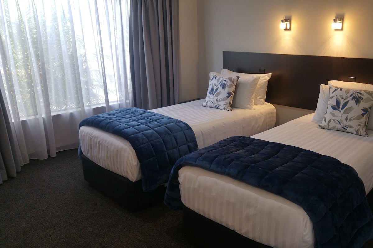 Habitación de hotel con dos camas, cada una con una colcha azul oscuro acolchada y ropa de cama blanca. Hay dos lámparas de pared sobre las camas y una ventana con cortinas translúcidas a la izquierda.