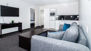 A modern living room with a grey sofa, a flat-screen TV mounted on the wall, a dark coffee table, and a kitchen area with white cabinets and a microwave. - White Heron Motor Lodge (Gisborne)