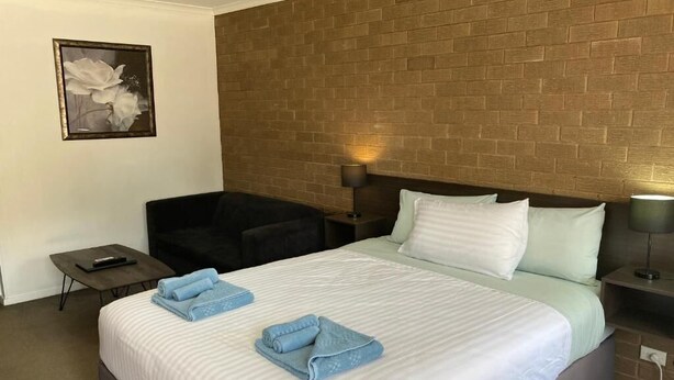 Una habitación de hotel con pared de ladrillo decorativa, una cama con sábanas blancas, dos almohadas, una silla, una mesita de noche con lámpara y un cuadro enmarcado en la pared.