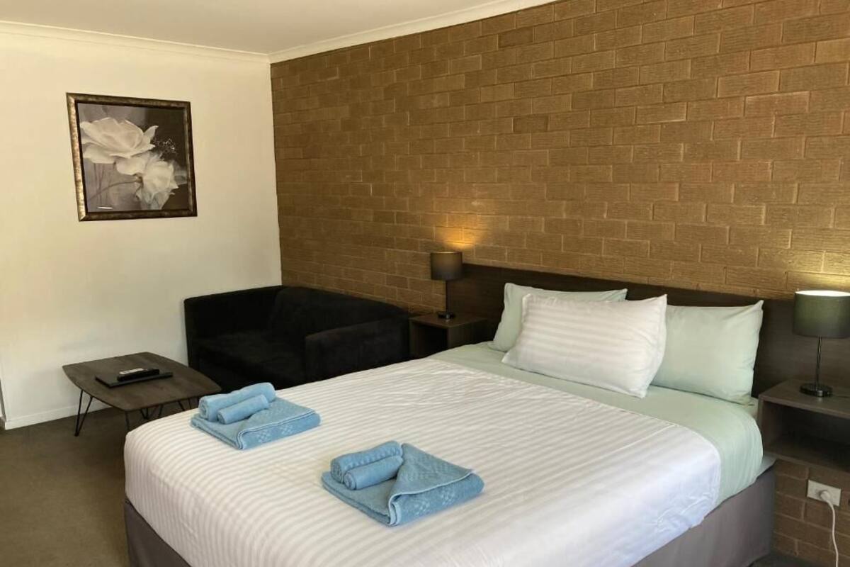 Ett hotellrum med en vägg som har en dekorativ tegelstensbeklädnad, en säng med vita sängkläder, två kuddar, en stol, ett nattduksbord med en lampa och en inramad tavla på väggen.