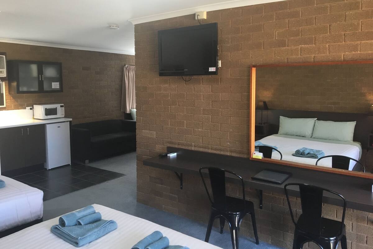 Ett hotellrum med ett litet kök, en säng med en spegel ovanför huvudändan, en TV som är monterad på väggen och ett skrivbord med stolar.