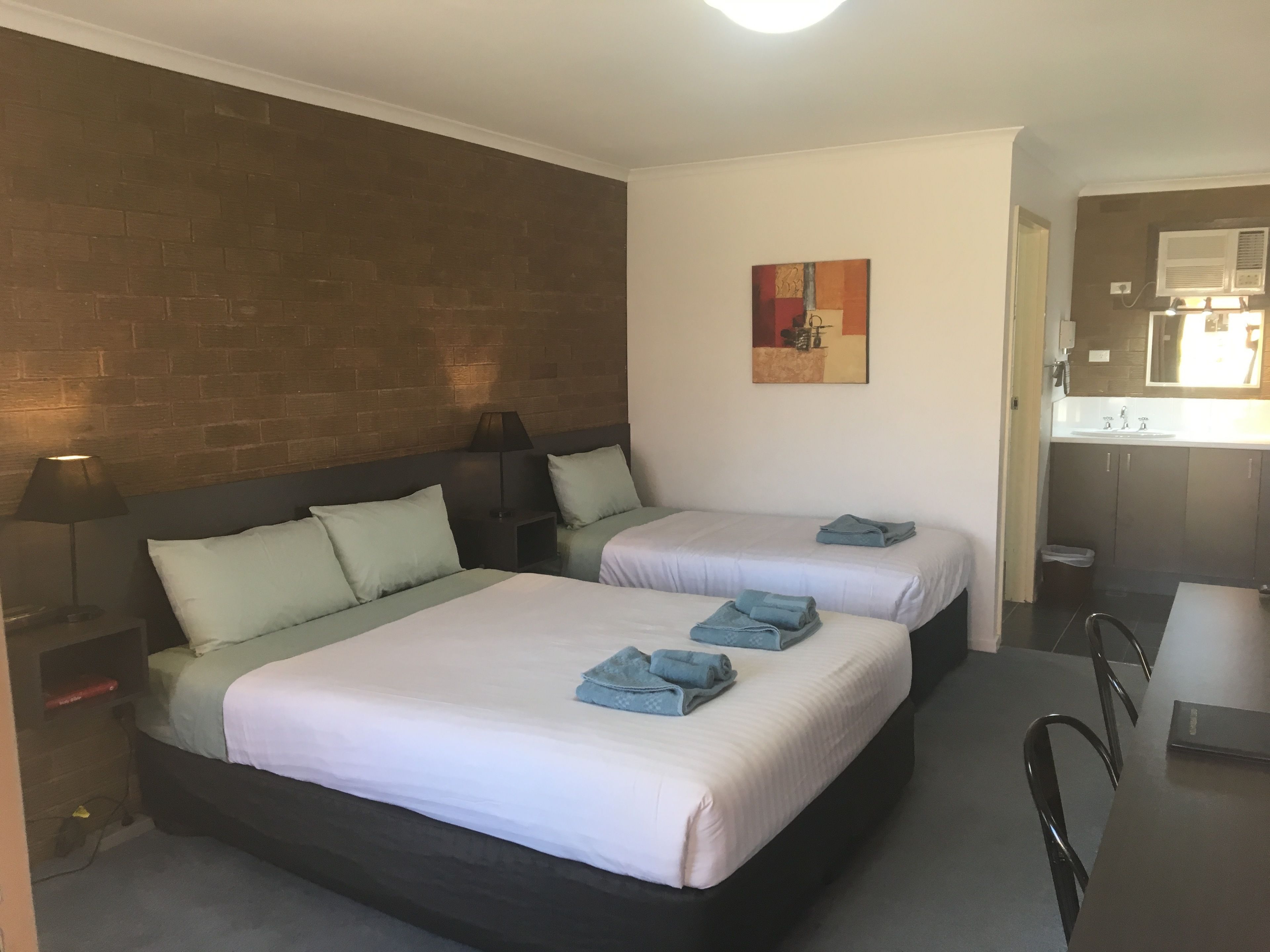 A hotel room with two beds, a small bedside table with a lamp, a painting on the wall, and a view of the bathroom.