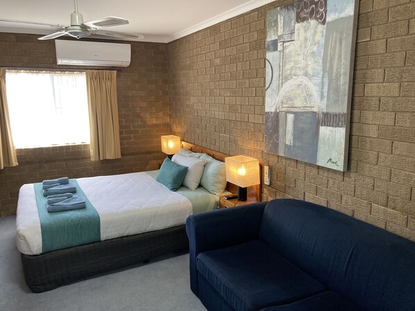 A hotel room with a bed, a blue armchair, a painting on the wall, and a ceiling fan.