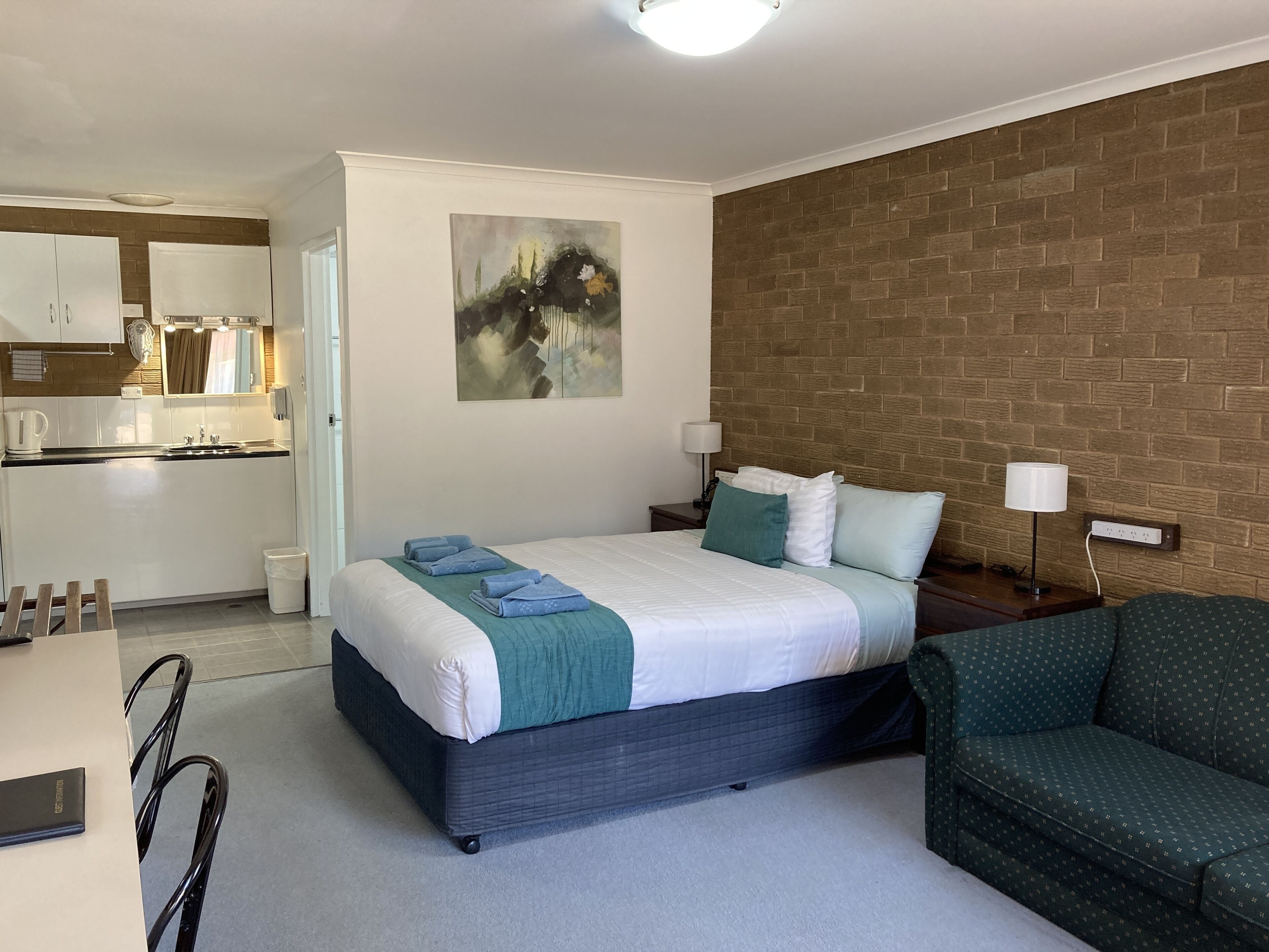 Una habitación de hotel con cama, mesitas de noche, un sillón verde, un cuadro en la pared y una cocina visible a través de una puerta abierta.