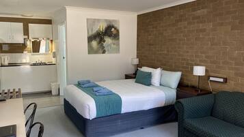 Una habitación de hotel con cama, mesitas de noche, un sillón verde, un cuadro en la pared y una cocina visible a través de una puerta abierta.