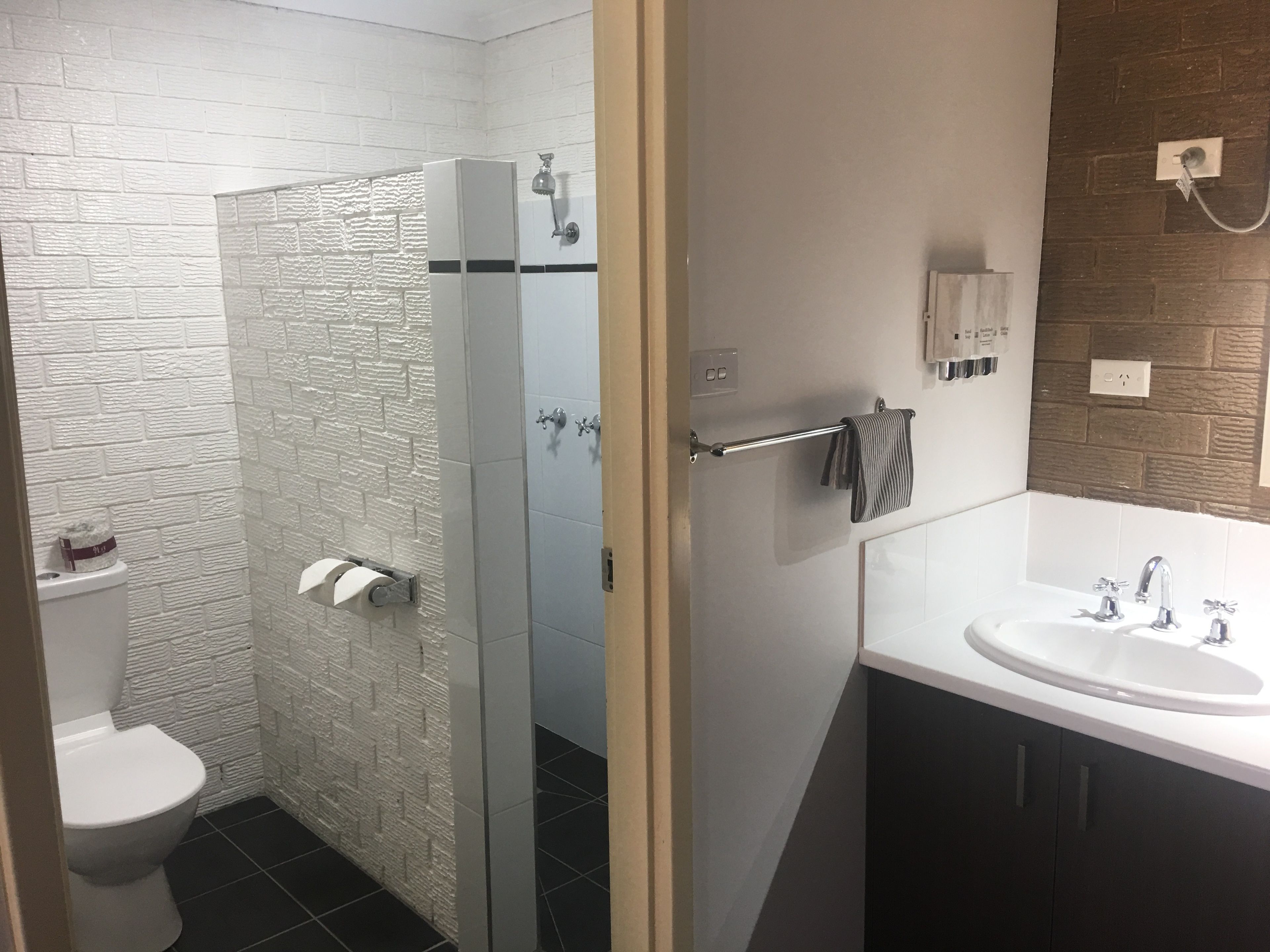 Un baño moderno con pared de ladrillo blanco, cabina de ducha de vidrio, inodoro blanco, lavamanos con espejo y toallero.