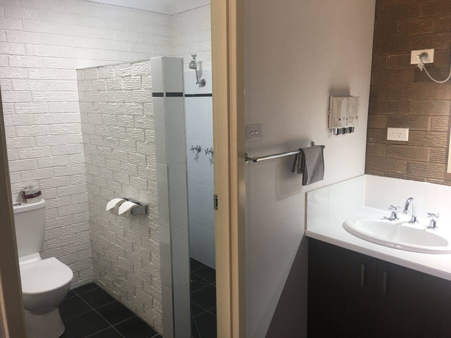 Un baño moderno con pared de ladrillo blanco, cabina de ducha de vidrio, inodoro blanco, lavamanos con espejo y toallero.