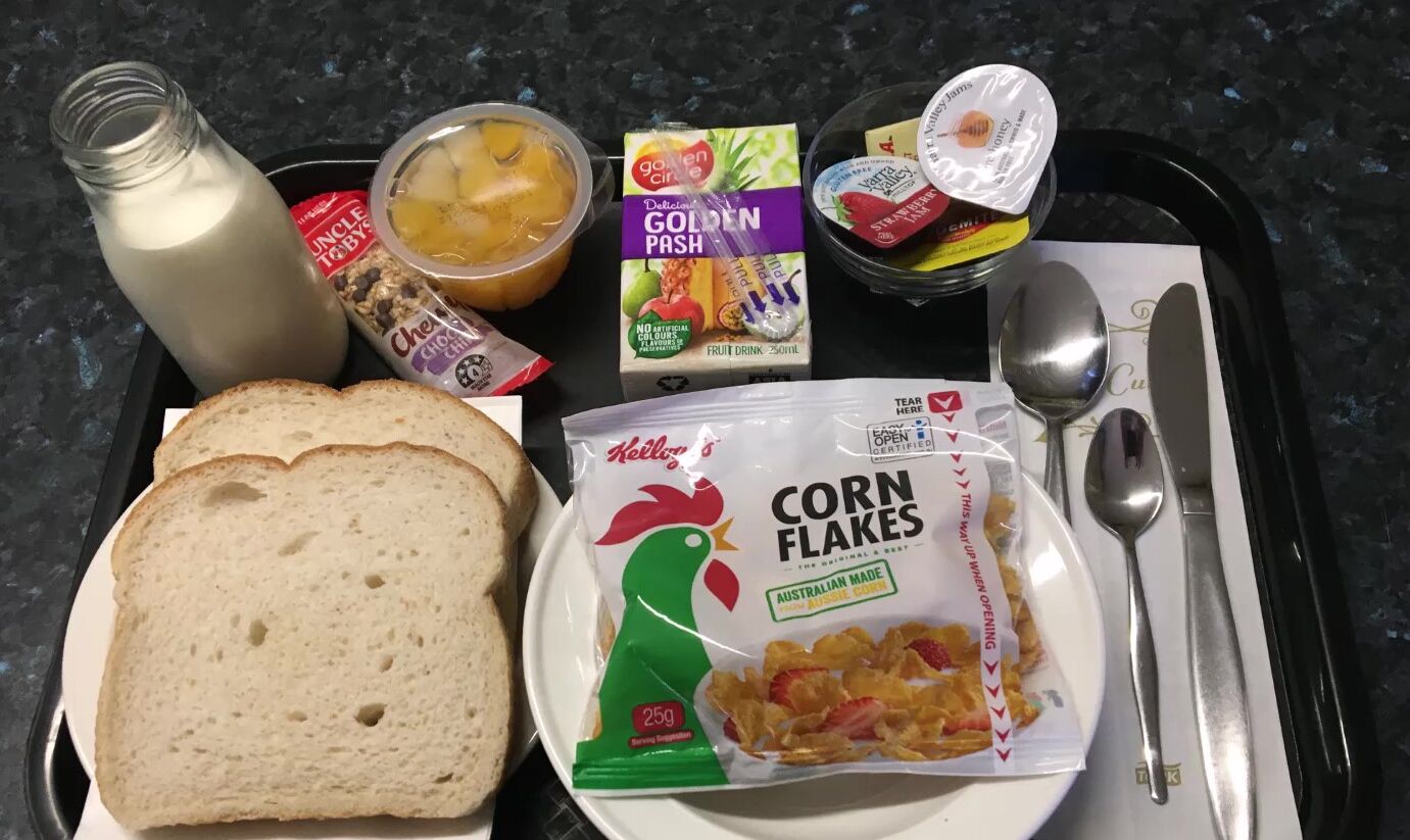 A tray with a bottle of milk, a bowl of fruit, a box of Golden Pash cereal, a packet of Kellogg's Corn Flakes, and slices of bread.