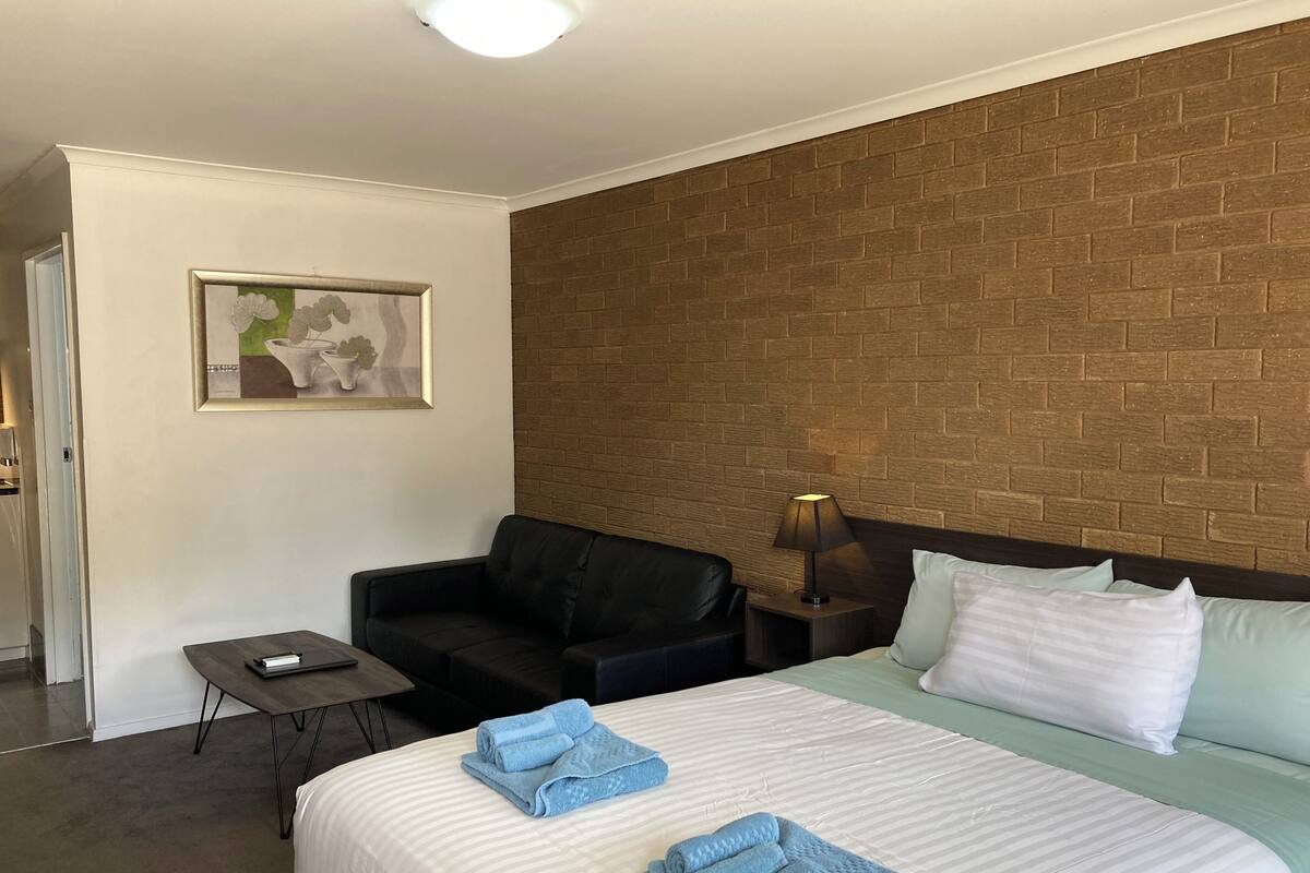Ett hotellrum med en säng, en svart soffa, ett litet bord, ett sängbord med en lampa och en vägg med ett mönster som efterliknar tegelstenar.