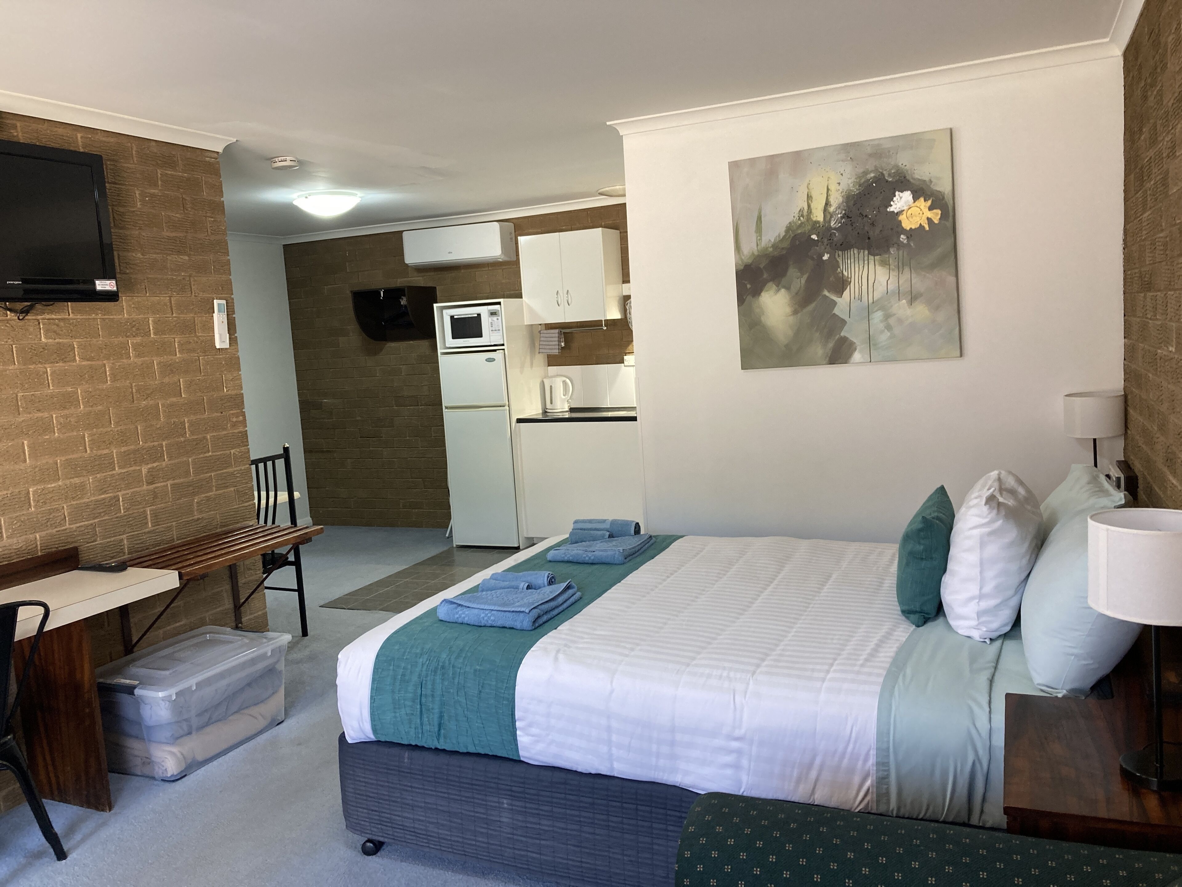 A hotel room with a bed, a desk, a chair, a TV, a microwave, and a kitchenette.