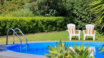 A swimming pool with two white chairs with orange cushions, surrounded by greenery.