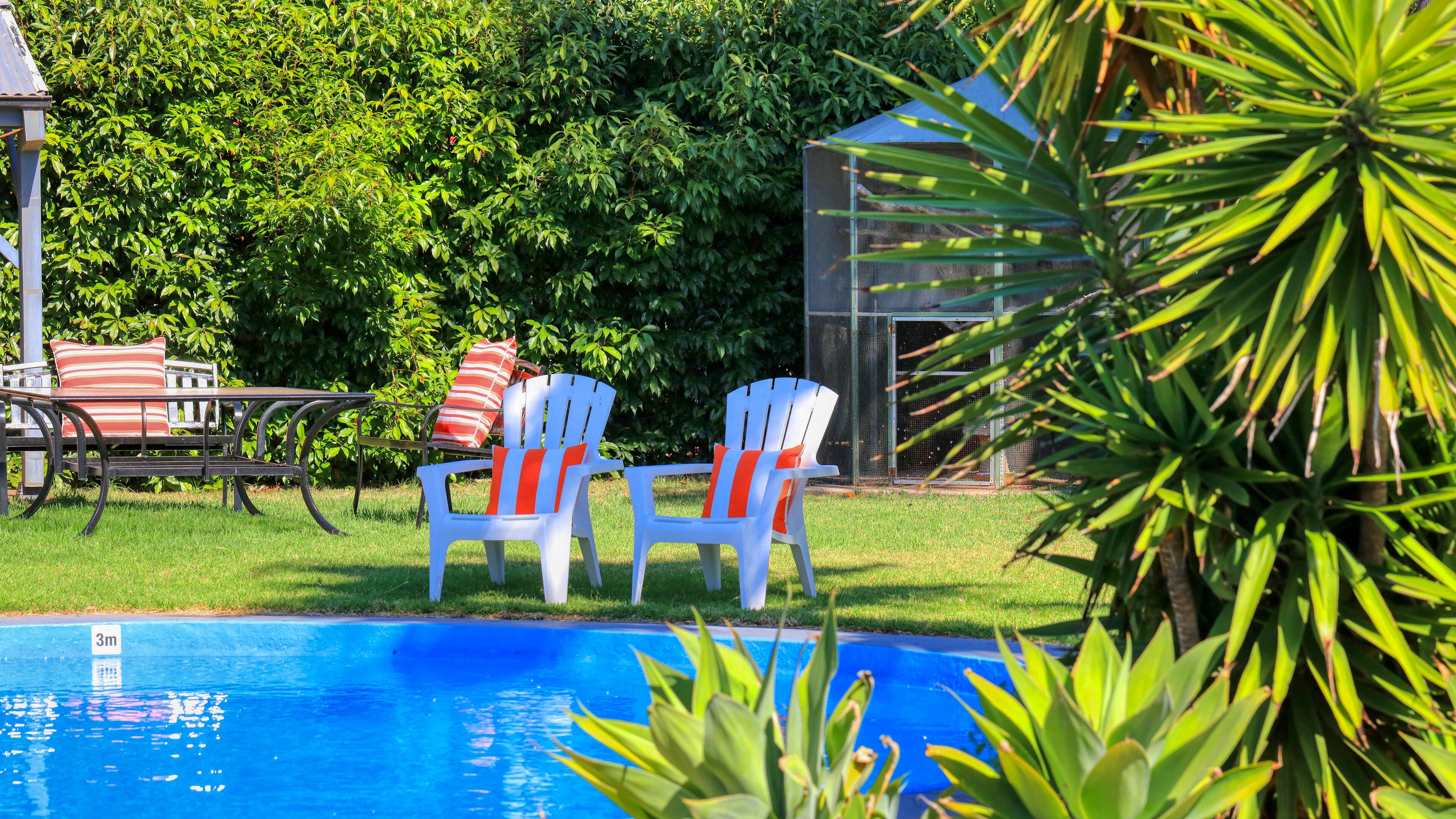 Un área junto a la piscina con dos sillas blancas con cojines anaranjados, una mesa con cojines a rayas y una piscina azul.