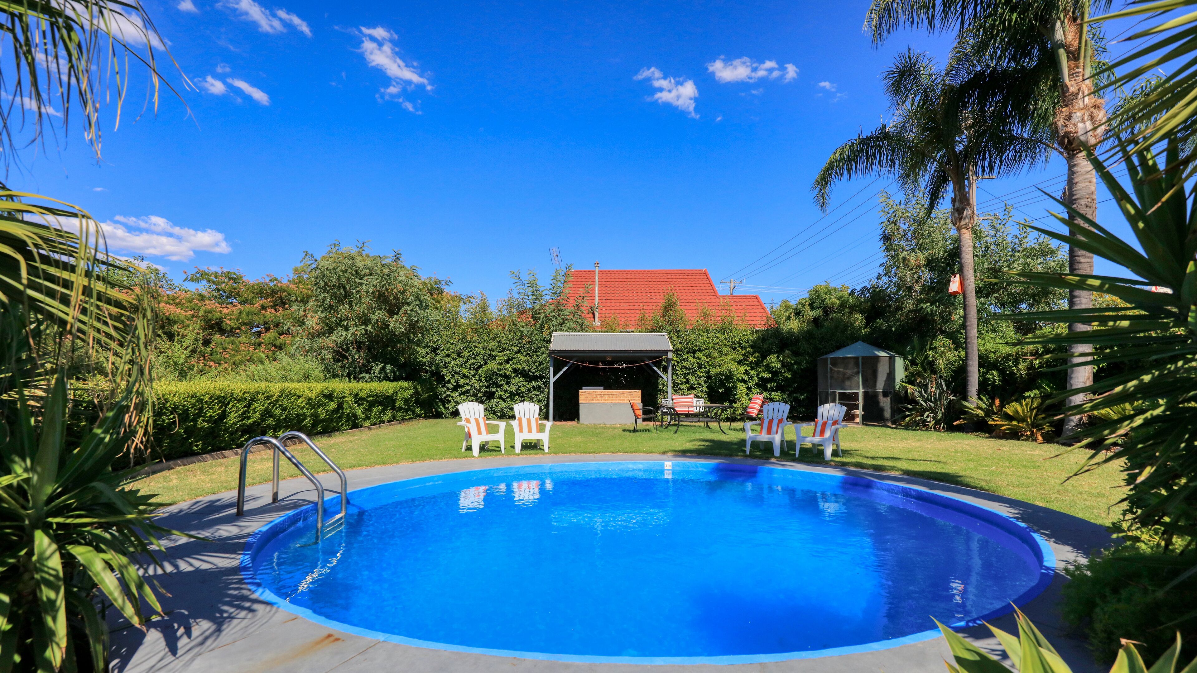 Una piscina con un área de terraza techada, rodeada de exuberante vegetación y un edificio con techo rojo al fondo.