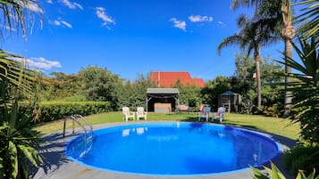 Una piscina con un área de terraza techada, rodeada de exuberante vegetación y un edificio con techo rojo al fondo.