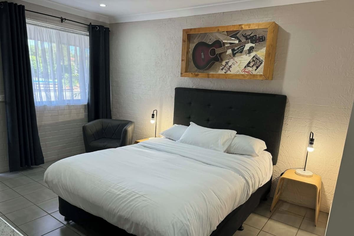 A bedroom with a large bed, a chair, a nightstand with a lamp, and a wall-mounted guitar display.