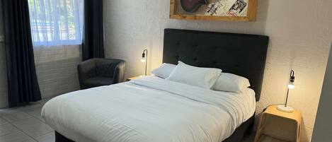 A bedroom with a large bed, a chair, a nightstand with a lamp, and a wall-mounted guitar display.