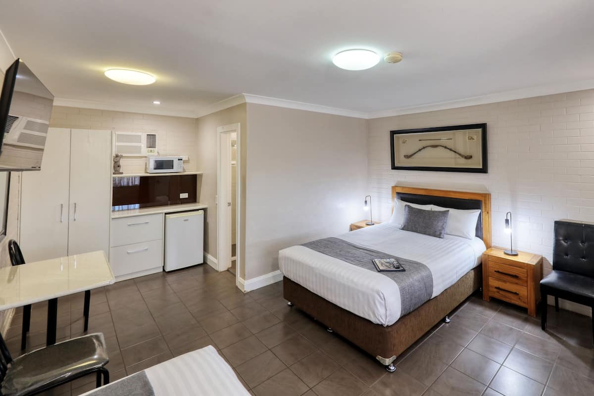 A modern hotel room with a bed, bedside table, chair, desk, kitchenette, and a framed picture on the wall.