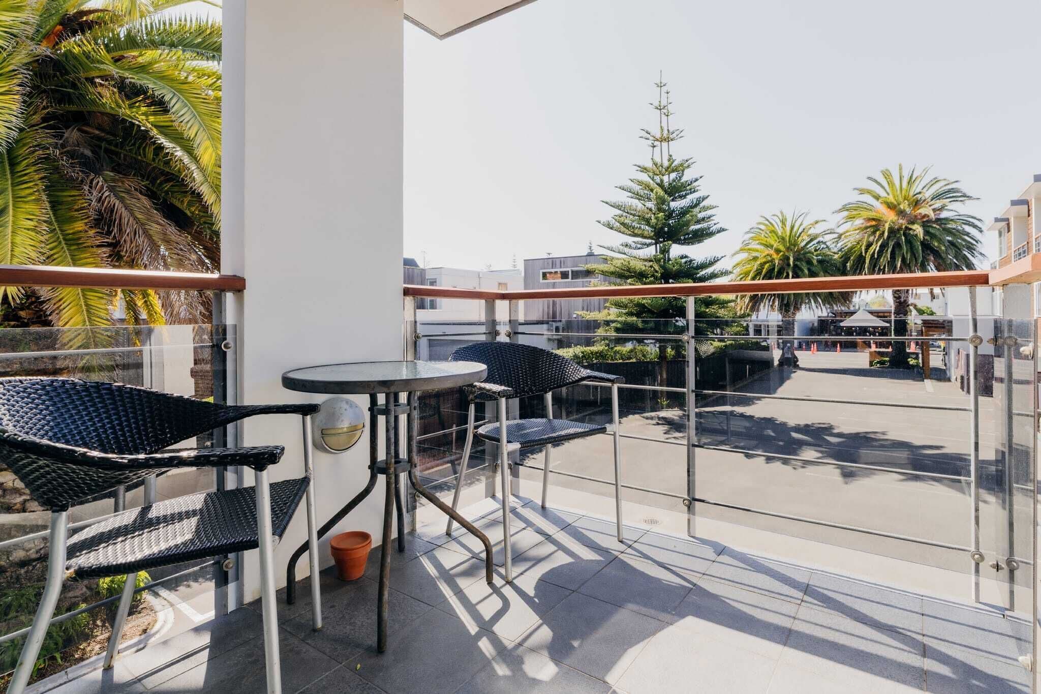 Un balcon avec deux fauteuils en osier noir et une petite table ronde, donnant sur une zone pavée agrémentée de palmiers et de bâtiments.