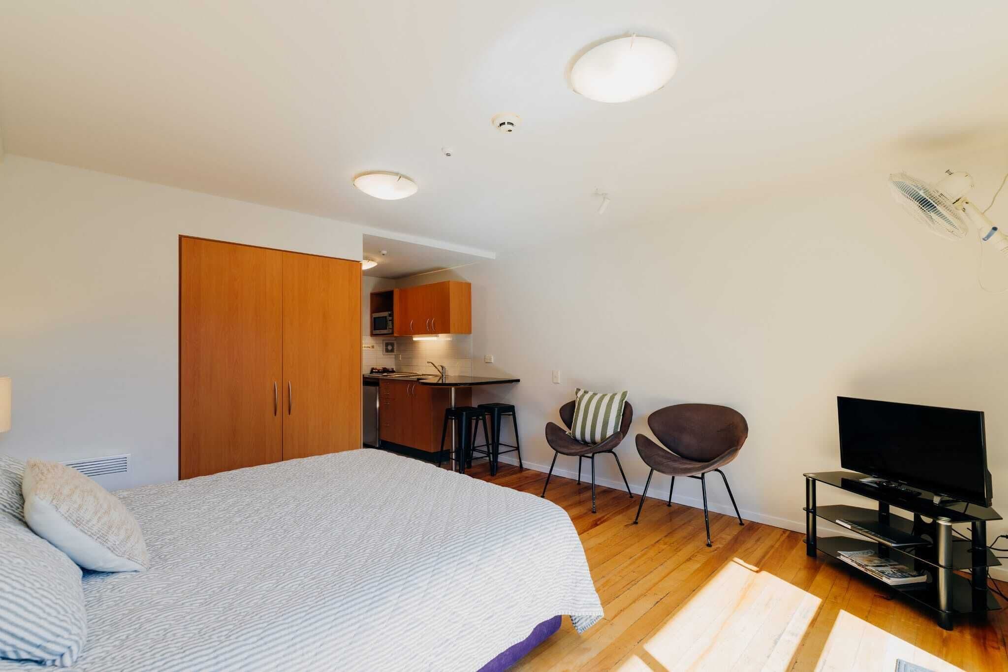 Un dormitorio con cama, televisor, una pequeña kitchenette y una zona de estar con sillas.