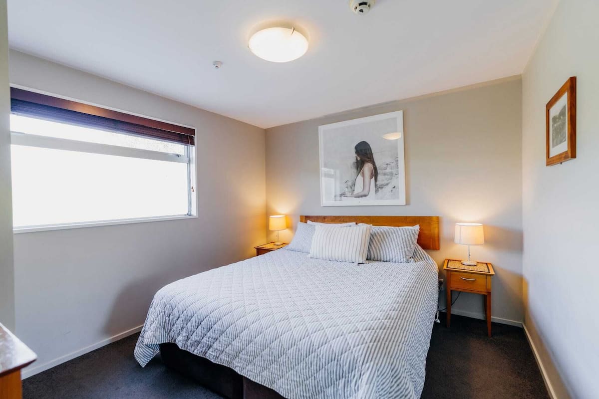 A neatly made bed with a quilted blanket, two bedside lamps, a framed artwork on the wall, and a window with blinds.