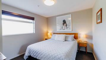 A neatly made bed with a quilted blanket, two bedside lamps, a framed artwork on the wall, and a window with blinds.