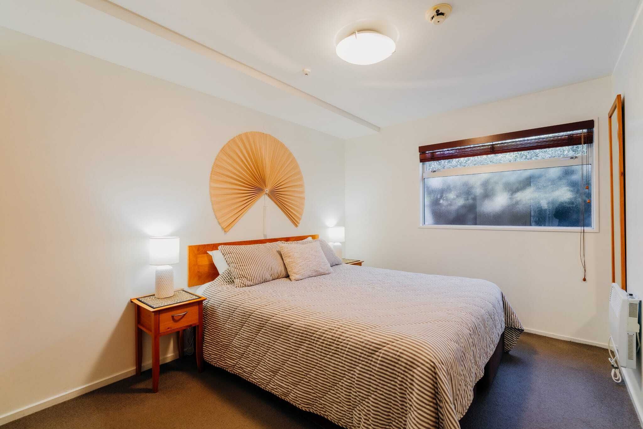 Un dormitorio con cama, lámparas de noche, ventilador en la pared, ventana con vistas y una lámpara de techo.