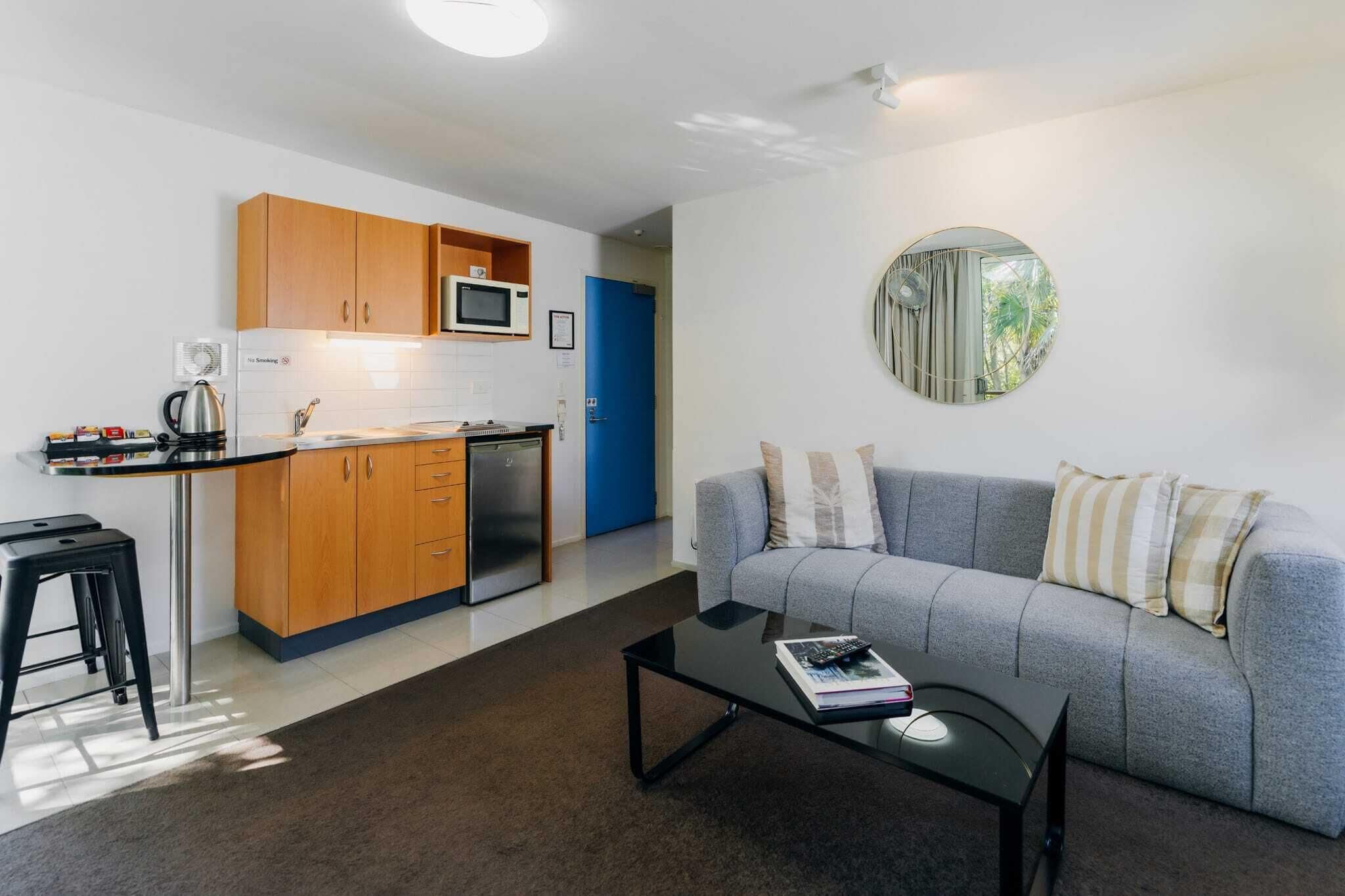 A compact living space with a kitchenette, a grey sofa, a coffee table, and a round mirror.