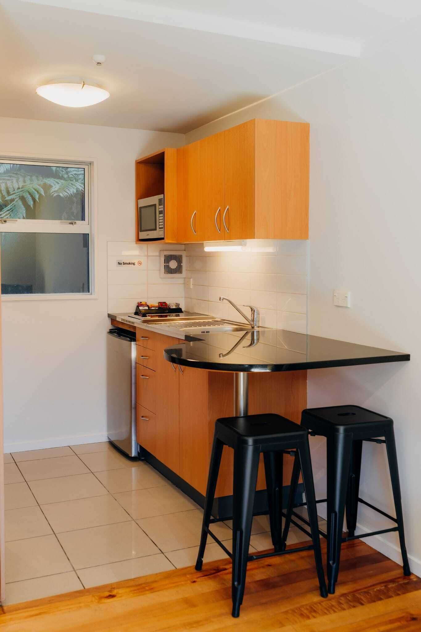 A compact kitchen with wooden cabinets, a microwave, a sink, and two black stools.