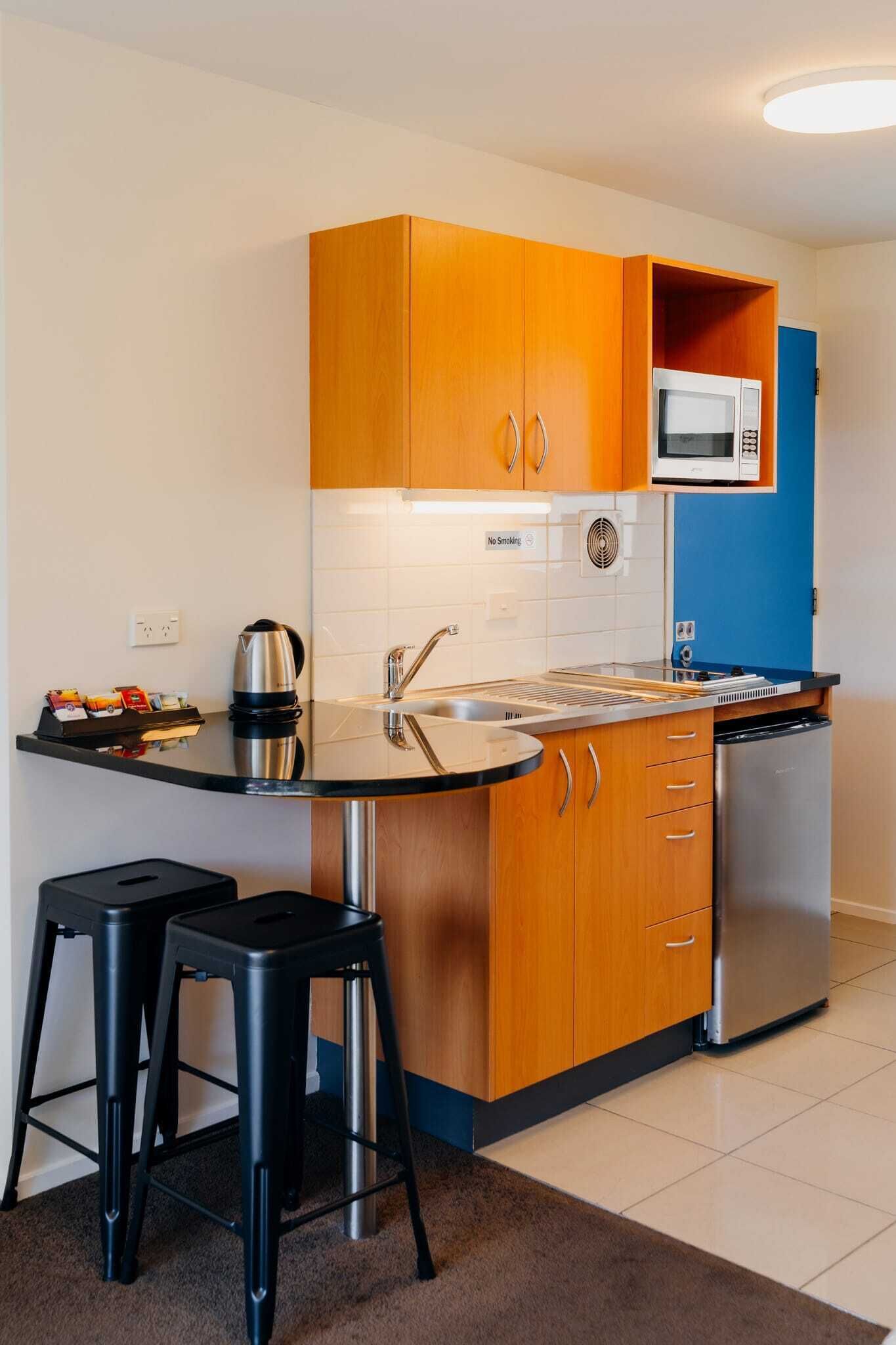A compact kitchen with wooden cabinets, a stainless steel sink, a microwave, and a dishwasher. There are two black bar stools and a kettle on the counter.