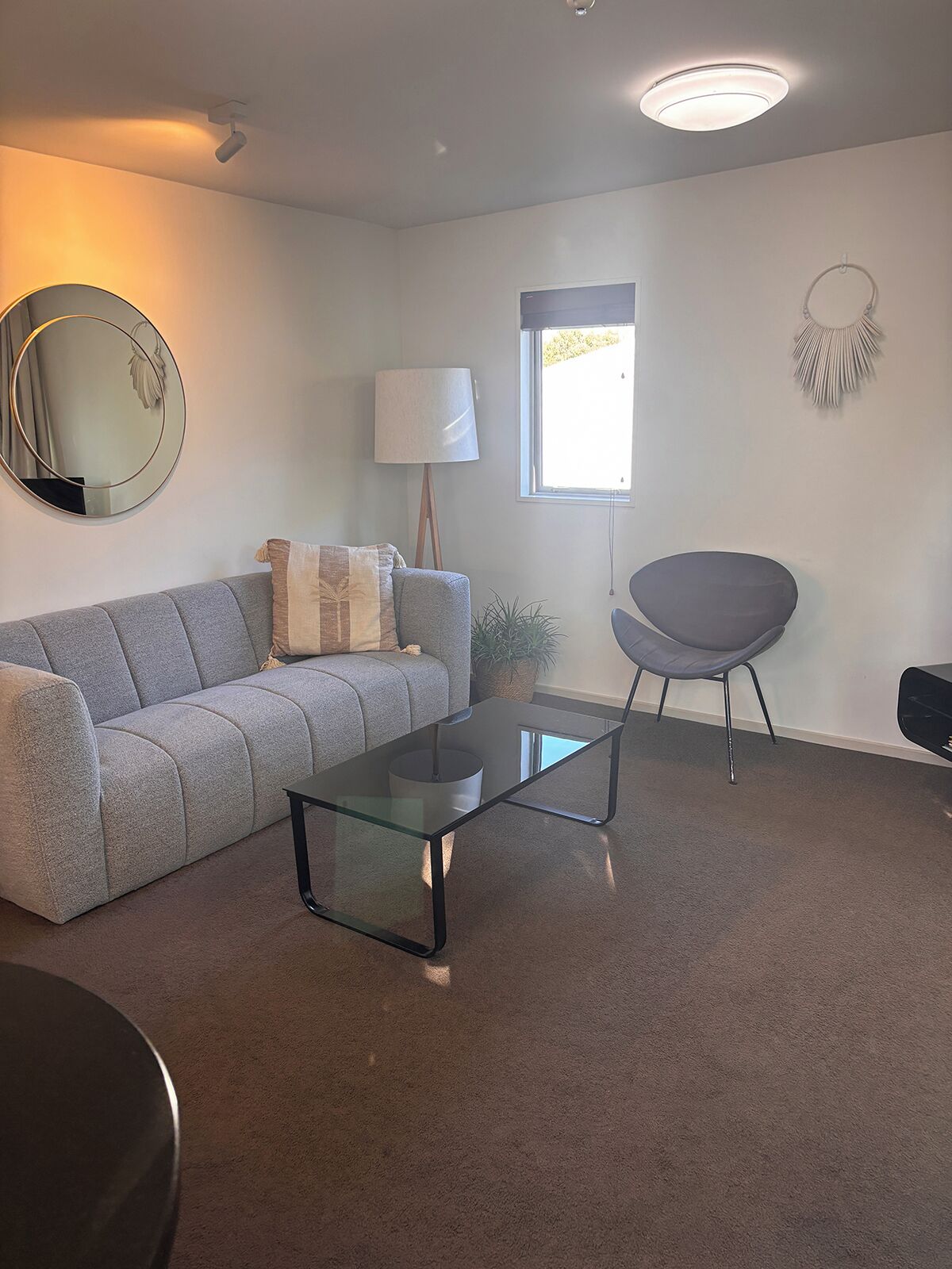A modern living room with a grey sofa, a glass coffee table, a grey armchair, a floor lamp, a wall-mounted mirror, a window, and a decorative wall piece.