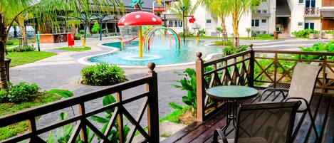 A pool area with a water slide and a mushroom-shaped structure, surrounded by greenery and a wooden deck with chairs.