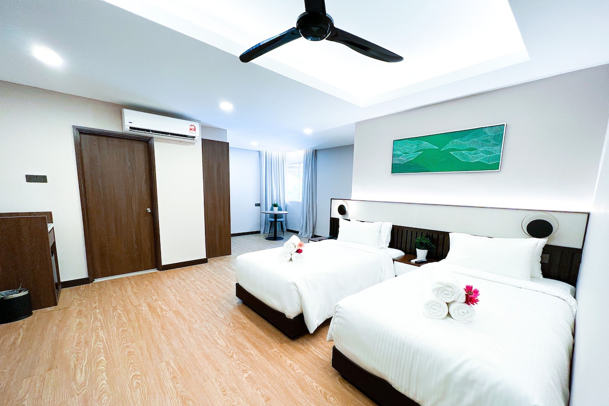 A hotel room with two beds, a ceiling fan, an air conditioning unit, a small table with a plant, and a painting on the wall.